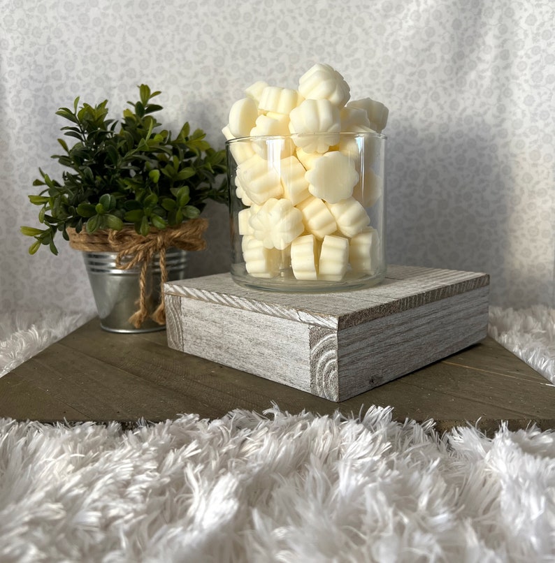 Puede incluir: Un tarro de cristal transparente lleno de ceras perfumadas de color marfil con forma de flor. El tarro est&aacute; sobre un bloque de madera blanqueada. A la izquierda hay una peque&ntilde;a planta en maceta en un cubo de metal galvanizado. El fondo es una tela con estampado floral.