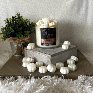 Puede incluir: Vela de aromaterapia en un tarro de vidrio con una etiqueta negra que dice "Relax" y "Black Chamomile". Derretidos de cera blanca est&aacute;n esparcidos alrededor del tarro sobre una superficie de madera. Una peque&ntilde;a planta en maceta est&aacute; en el fondo.