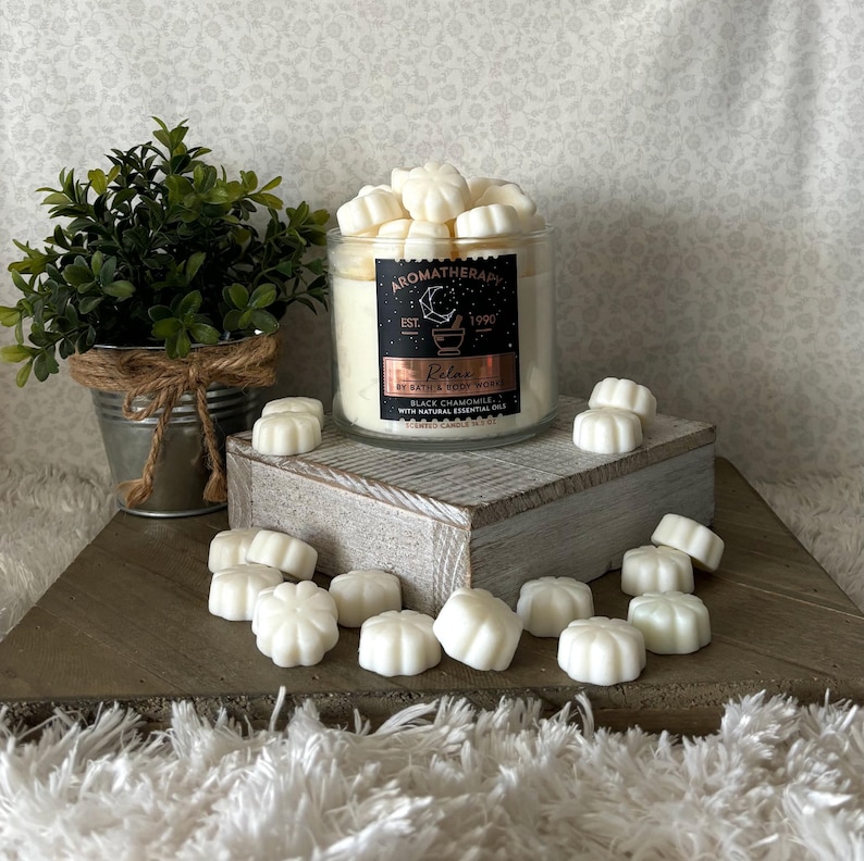 Puede incluir: Vela de aromaterapia en un tarro de vidrio con una etiqueta negra que dice "Relax" y "Black Chamomile". Derretidos de cera blanca est&aacute;n esparcidos alrededor del tarro sobre una superficie de madera. Una peque&ntilde;a planta en maceta est&aacute; en el fondo.