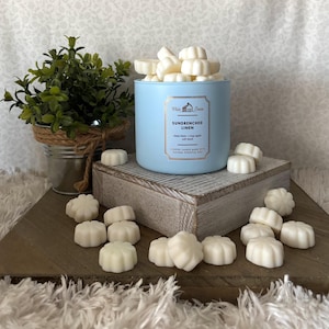 May include: A blue candle jar with a white lid filled with white wax melts. The jar has the text "White Barn Sundrenched Linen" and "Clean Linen - Crisp Apple - Soft Musk" on the front. The wax melts are shaped like flowers and are scattered around the jar on a wooden surface.