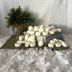 Puede incluir: Una colecci&oacute;n de ceras perfumadas en forma de flor de color marfil. Las ceras est&aacute;n dispuestas sobre una caja de madera r&uacute;stica y esparcidas sobre una superficie blanca y esponjosa. A la izquierda hay una peque&ntilde;a planta en maceta en un cubo de metal galvanizado.