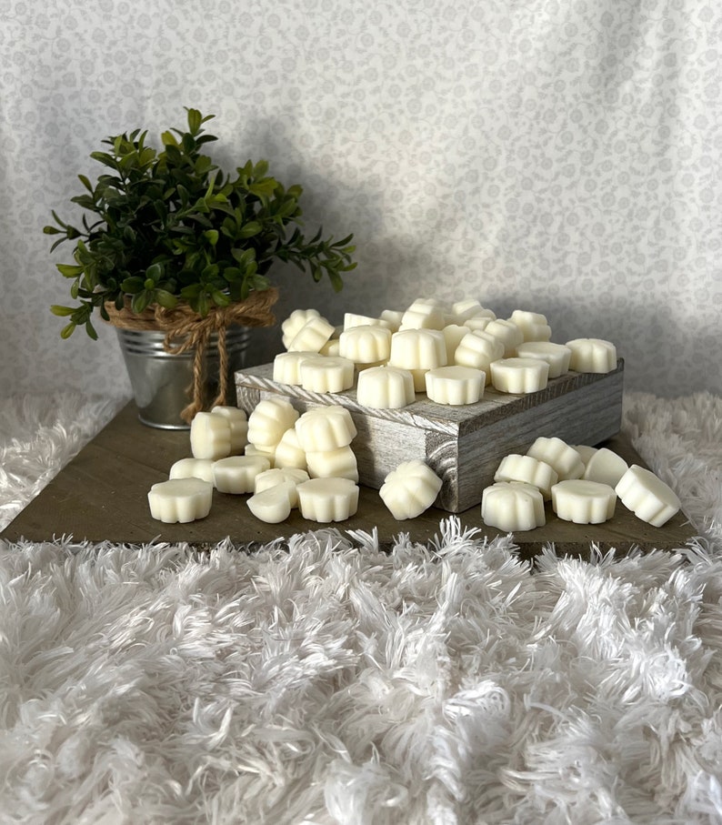 Puede incluir: Una colecci&oacute;n de ceras perfumadas en forma de flor de color marfil. Las ceras est&aacute;n dispuestas sobre una caja de madera r&uacute;stica y esparcidas sobre una superficie blanca y esponjosa. A la izquierda hay una peque&ntilde;a planta en maceta en un cubo de metal galvanizado.