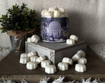 Può includere: Una candela in barattolo di vetro con un'etichetta con una scena invernale, piena di cere profumate bianche. L'etichetta recita "Lavender & Pine". Le cere sono sparse su una superficie di legno. Una piccola pianta in vaso è sullo sfondo.