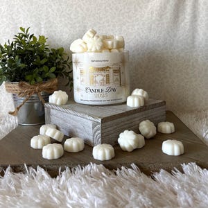 May include: A white candle jar with gold accents and the text "Candle Day 2025" is surrounded by small, white, flower-shaped wax melts. A small potted plant sits to the left of the candle on a wooden surface.
