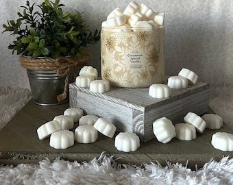 Może przedstawiać: Szklany słoik wypełniony białymi woskowymi kostkami, oznaczony etykietą "Cinnamon Spiced Vanilla". Słoik otoczony jest dodatkowymi woskowymi kostkami na drewnianej powierzchni.