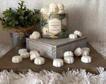 May include: A Bath & Body Works Morning Tea soy wax blend candle in a green jar with floral and tea cup illustrations, surrounded by small white flower-shaped wax melts, with a green potted plant in a metal bucket nearby.