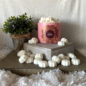 May include: A pink candle with gold accents and the text "Danbury Shortbread" sits on a wooden block, surrounded by white wax melts. A small potted plant in a metal bucket is on the left.
