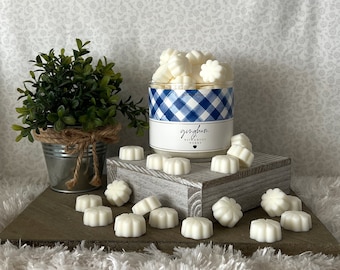 Può includere: Un barattolo bianco con un motivo a quadri blu e bianchi riempito di cere profumate bianche. Il barattolo è etichettato "gingham" e "Bath & Body Works". Ci sono diversi cere profumate bianche sparse su una superficie di legno.