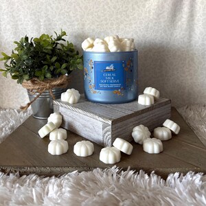 May include: A light blue candle with the text "Cereal Milk Soft Serve" and white wax melts. The candle sits on a wooden block with scattered wax melts. A small potted plant is in the background.