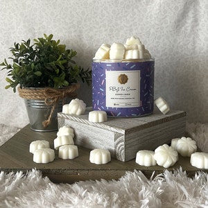 May include: A cylindrical candle jar with a purple background and the text "PB&J Ice Cream". The jar is filled with white wax melts, and more are scattered around a wooden platform. A small potted plant sits to the left.