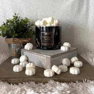 May include: A black and white marbled glass candle jar with the text "Vanilla Noir" and white wax melts. The candle is on a wooden stand with a small potted plant in a metal bucket. The wax melts are scattered around the stand.