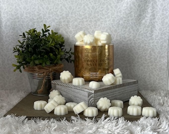 May include: A gold-colored candle holder with the text "FORCE OF NATURE" and white wax melts in a floral shape. A small potted plant sits to the left. The melts are scattered on a wooden block and a white, fluffy surface.