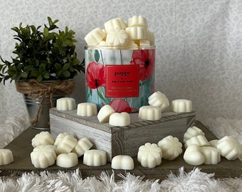 May include: White wax melts in the shape of flowers and hearts, arranged on a wooden surface. The wax melts are next to a glass jar with a floral design and the text "poppy bath & body works" on the label.