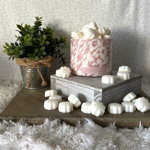 May include: A pink and white swirl candle jar with the text "Hot Pink Cocoa" and "Scented Candle" on the label. The candle is surrounded by white wax melts shaped like flowers.