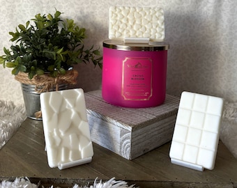 Può includere: Tre cubetti di cera bianca, ognuno con un motivo diverso, sono esposti su una superficie di legno. I cubetti di cera sono confezionati in una pellicola di plastica trasparente. I cubetti di cera sono accanto a un barattolo di candela rosa con un coperchio dorato. Il barattolo di candela ha un'etichetta che recita "Cactus Blossom".