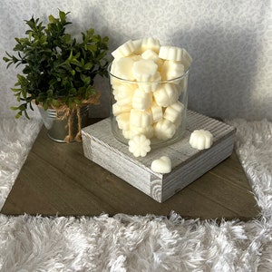 Puede incluir: Un tarro de cristal transparente lleno de ceras perfumadas de color marfil en varias formas florales. El tarro est&aacute; sobre un bloque de madera blanco envejecido, con una peque&ntilde;a planta en maceta en el fondo. El arreglo est&aacute; sobre una superficie de madera en forma de espiga.