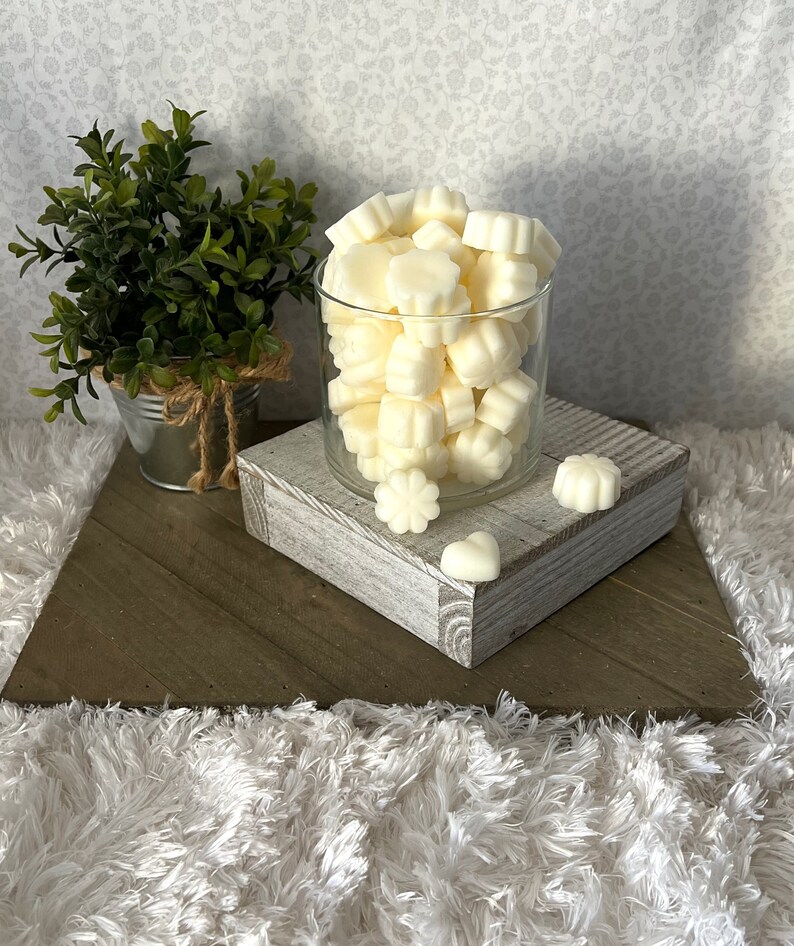 Puede incluir: Un tarro de cristal transparente lleno de ceras perfumadas de color marfil en varias formas florales. El tarro est&aacute; sobre un bloque de madera blanco envejecido, con una peque&ntilde;a planta en maceta en el fondo. El arreglo est&aacute; sobre una superficie de madera en forma de espiga.