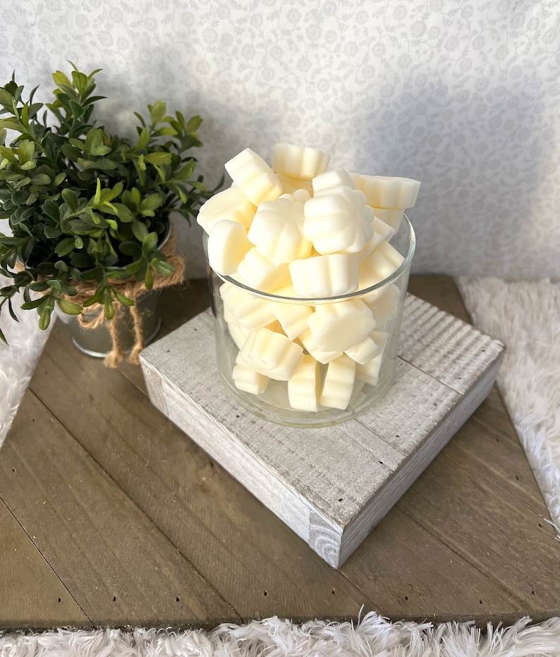 Puede incluir: Un tarro de cristal transparente lleno de ceras perfumadas de color marfil con forma de peque&ntilde;as flores. El tarro se encuentra sobre un bloque de madera blanco y gris, que a su vez est&aacute; colocado sobre una superficie de madera m&aacute;s grande y en &aacute;ngulo. Una peque&ntilde;a planta en maceta es visible en el fondo.