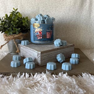 May include: A glass jar filled with light blue wax melts, alongside a blue-labeled candle with popcorn and slushie graphics. The candle sits on a wooden block, with additional wax melts scattered around. A small potted plant is in the background.