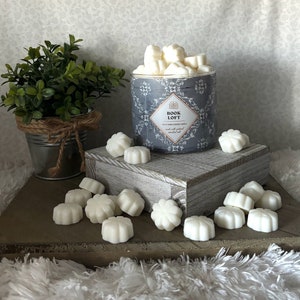 May include: A gray candle jar with the text "BOOK LOFT" and a floral design. The jar is filled with white wax melts shaped like flowers. The wax melts are scattered on a wooden surface.