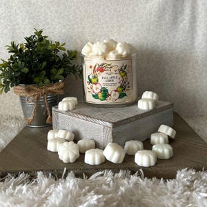 May include: A glass jar filled with white wax melts shaped like flowers. The jar has a label that says "Fall Apple Cider" with a picture of apples and leaves. The wax melts are sitting on a white wooden surface.
