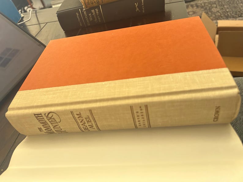 May include: A hardcover book titled "The Mammoth Hunters" by Jean M. Auel. The book has an orange cover and a cream-colored spine with the author's name and the publisher's name, "Crown."