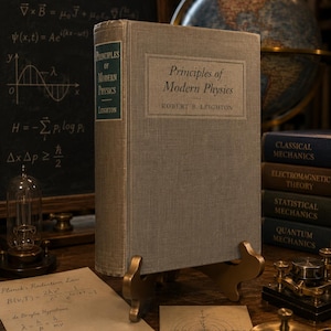 May include: A hardback book titled Principles of Modern Physics by Robert B. Leighton with a light grey fabric cover, displayed on a gold-toned metal stand. The spine features a dark green label with white text reading Principles of Modern Physics.
