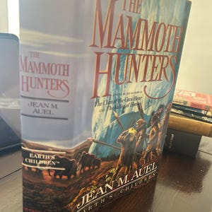 May include: A hardcover book titled "The Mammoth Hunters" by Jean M. Auel. The cover features a landscape with mammoths and figures, with the title in large, stylized letters. The book is part of the "Earth's Children" series.
