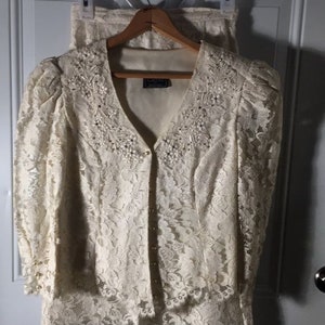 May include: A white lace jacket and skirt set with beaded embellishments. The jacket has a V-neckline and long sleeves. The skirt is knee-length and has a scalloped hem.