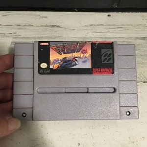 May include: A gray Super Nintendo Entertainment System game cartridge with a colorful label featuring the text "Aero the Acrobatic Squirrel" and a cartoon squirrel in a plane. The label also includes the text "Super Nintendo" and "Nintendo of America Inc."