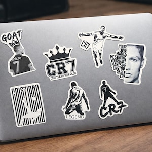 Puede incluir: Un port&aacute;til cubierto de pegatinas en blanco y negro con Cristiano Ronaldo. Las pegatinas incluyen im&aacute;genes de Ronaldo en acci&oacute;n, su logotipo CR7 y texto como "GOAT", "LEGEND" y una cita sobre el amor y el odio.
