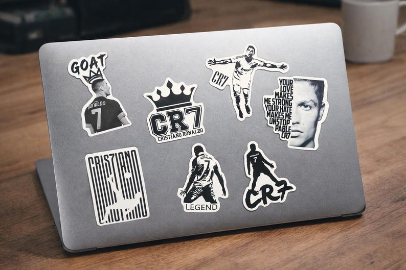 Puede incluir: Un port&aacute;til cubierto de pegatinas en blanco y negro con Cristiano Ronaldo. Las pegatinas incluyen im&aacute;genes de Ronaldo en acci&oacute;n, su logotipo CR7 y texto como "GOAT", "LEGEND" y una cita sobre el amor y el odio.