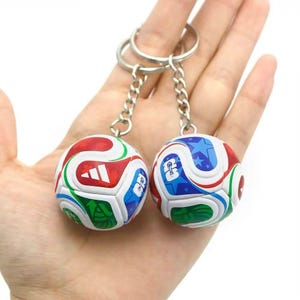 Puede incluir: Dos llaveros de balón de fútbol. Cada balón es una réplica en miniatura con paneles rojos, blancos, azules y verdes. Los llaveros están unidos por una cadena y un anillo plateados. Los balones tienen un logotipo y un diseño de estrellas.