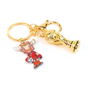 May include: A gold-toned metal keychain featuring a circular ring attached to a lobster clasp. The keychain includes two charms: a three-dimensional gold-colored trophy replica and a flat, colorful enamel charm of a cartoon moose playing soccer.