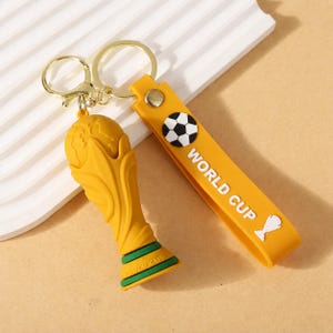 May include: A gold-colored keychain featuring a trophy and a yellow strap with "WORLD CUP" text and a soccer ball graphic. The trophy has a globe design. The keychain has a gold-colored clasp.