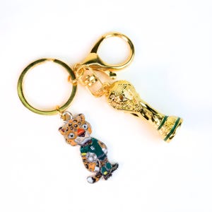 May include: A gold-toned metal keychain featuring two charms: a three-dimensional trophy cup and a flat enamel charm depicting a stylized leopard wearing a green jersey and holding a soccer ball, attached to a circular ring and a lobster clasp.
