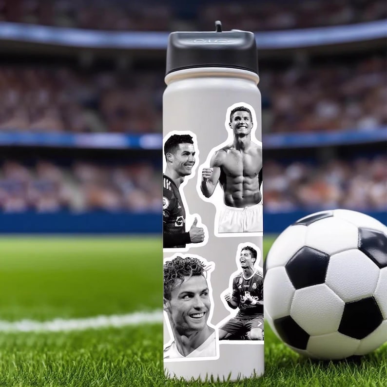 Puede incluir: Una botella de agua blanca con tapa negra, adornada con pegatinas en blanco y negro de un futbolista. Un bal&oacute;n de f&uacute;tbol descansa sobre la hierba verde junto a la botella. El fondo muestra un estadio borroso.