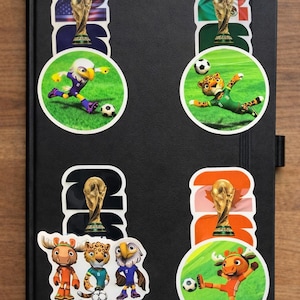 Pu&ograve; includere: Un taccuino nero con adesivi raffiguranti mascotte di cartoni animati e il trofeo della Coppa del Mondo FIFA. Gli adesivi presentano un'aquila, un giaguaro, un alce e un uccello, ognuno che gioca a calcio. Gli adesivi includono l'anno 2022 e bandiere.