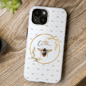 May include: White iPhone case with a gold crown and bee design. The case has a white background with a repeating pattern of gold crowns. The bee is surrounded by a gold floral design and the word "Queen" is written in gold lettering.