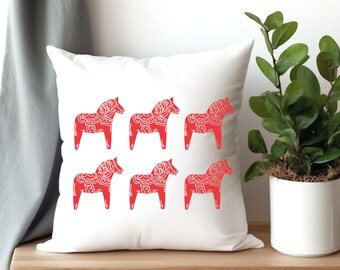 May include: White square throw pillow featuring six red Dala horse designs. The horses are arranged in two rows of three, with intricate patterns within their silhouettes. The pillow is placed on a wooden surface, with a plant in a white pot to the right.