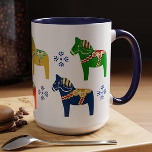 May include: White ceramic mug with a blue rim and handle. The mug features a pattern of colorful Swedish Dala horses with snowflakes.