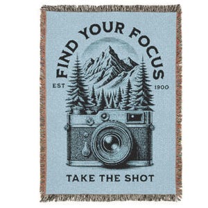 May include: A light blue woven throw blanket with fringed edges featuring a black graphic illustration of a vintage camera, pine trees, and a mountain range. The text reads FIND YOUR FOCUS, EST 1900, and TAKE THE SHOT in a bold black font.