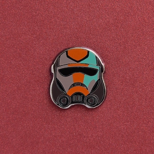 Op de afbeelding: Een kleurrijke emaille pin met een gestileerde Stormtrooper helm. Het helmontwerp bevat oranje, turquoise en grijze accenten, met een zwart vizier en een zilveren rand. De pin is geplaatst tegen een rode achtergrond.