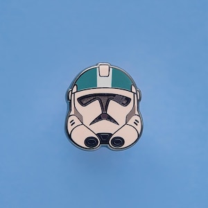 May include: A pin featuring a white and black helmet design with a teal stripe and accents. The helmet has a distinctive visor and is set against a solid blue background. The pin is likely a collectible or accessory.