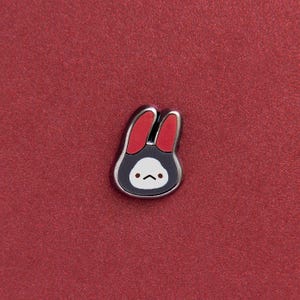 Puede incluir: Un pin de conejo negro y plateado con orejas rojas y una cara blanca con una boca pequeña y curvada. El pin está colocado sobre un fondo rojo texturizado.