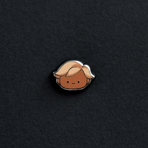 Op de afbeelding: Een kleine emaille pin met een cartoonfiguur met een bruine huid, blond haar en een glimlachend gezicht.