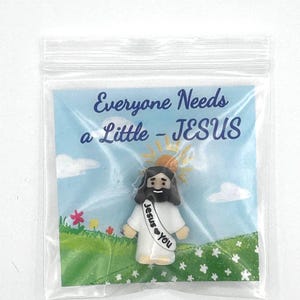 May include: A small figurine of Jesus in a plastic bag. The figurine has a white robe and a banner that says "Jesus ♥ You". The background is a blue sky with the text "Everyone Needs a Little - JESUS".