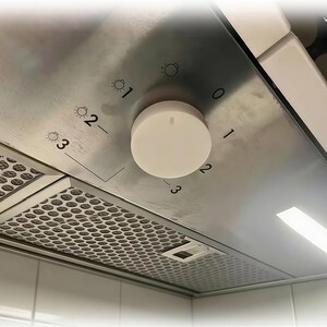 May include: A stainless steel range hood control panel featuring a circular white knob. The panel displays numerical settings from 0 to 3 and light intensity icons labeled 1, 2, and 3. A metallic mesh grease filter is visible on the underside.
