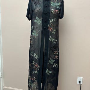 May include: A sheer black knee-length duster featuring short sleeves, a mandarin collar, and a front closure with fabric knot buttons. The garment is decorated with embroidered dragonfly motifs in shades of green, brown, and black throughout.