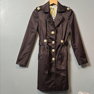 May include: A dark brown trench coat featuring a wide collar, long sleeves, and a waist belt. The coat is adorned with gold-toned buttons on the shoulders and down the front closure. The interior lining displays a multi-colored abstract pattern.